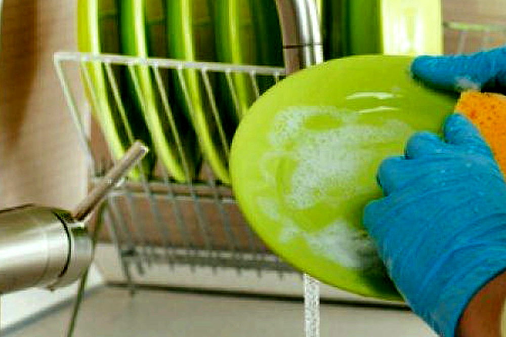 A gloved hand washing a plate at a kitchen sink
